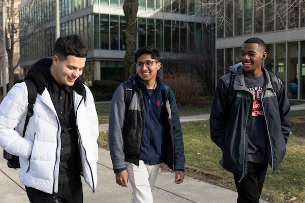 Three male students walk down a sidewalk and laugh together while carrying backpacks and wearing coats.