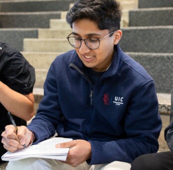 Young male Indian student takes notes in his notebook while sitting on a staircase.
                  