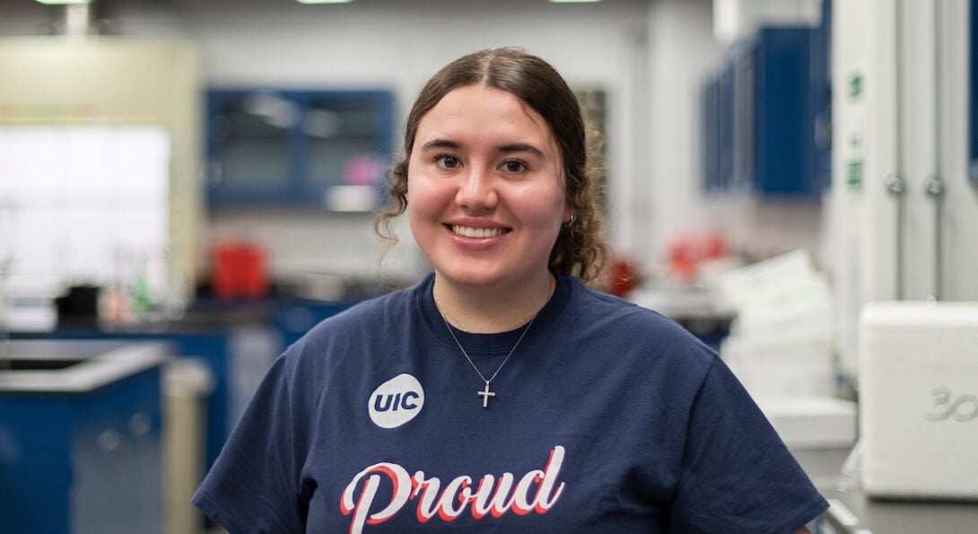 Young female student with curly brown hair stands smiling in a lab. She is wearing a blue t shirt with the UIC logo and the word proud on the front.