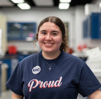 Young female student with curly brown hair stands smiling in a lab. She is wearing a blue t shirt with the UIC logo and the word proud on the front.
                  