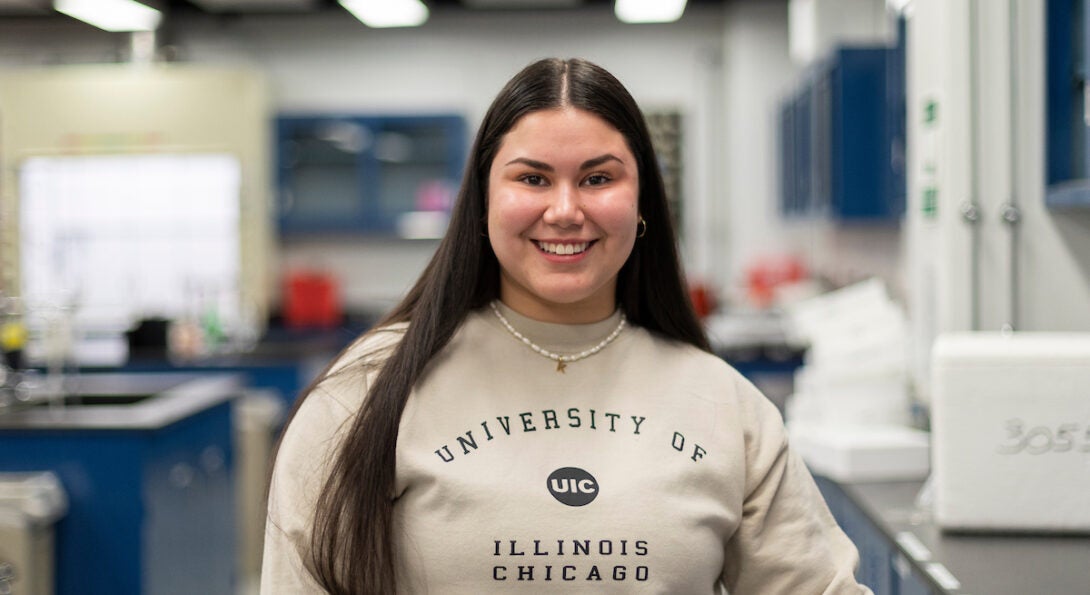 Biological Sciences student with long, straight brown hair smiles in a lab. She is wearing a tan sweatshirt with the University of Illinois Chicago logo on it.