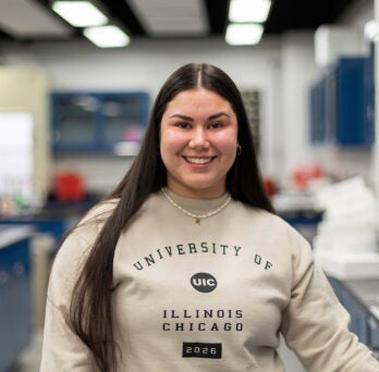 Biological Sciences student with long, straight brown hair smiles in a lab. She is wearing a tan sweatshirt with the University of Illinois Chicago logo on it.
                  