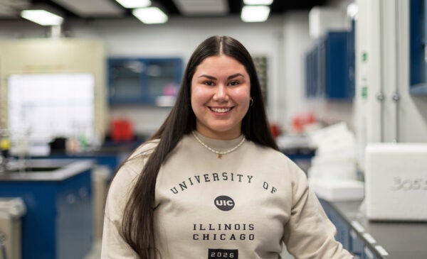 Biological Sciences student with long, straight brown hair smiles in a lab. She is wearing a tan sweatshirt with the University of Illinois Chicago logo on it.