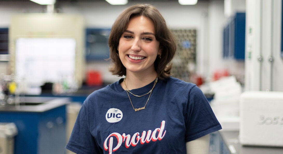 College of Liberal Arts and Sciences student Daytonna smiles in a lab. She is a young white woman with short brown hair, wearing a navy blue short sleeve shirt that says UIC Proud.