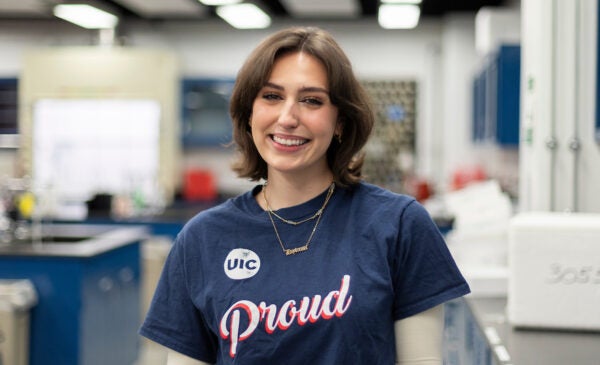 College of Liberal Arts and Sciences student Daytonna smiles in a lab. She is a young white woman with short brown hair, wearing a navy blue short sleeve shirt that says UIC Proud.