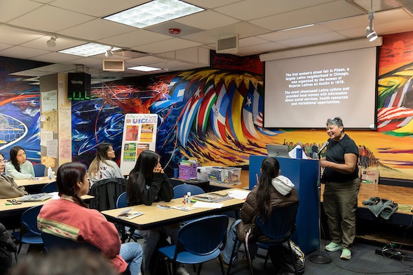 Local Chicago artist Diana Solis speaks at the Latino Cultural Center on the University of Illinois Chicago's east campus. The room is covered in murals and students sit at tables while Diana presents.