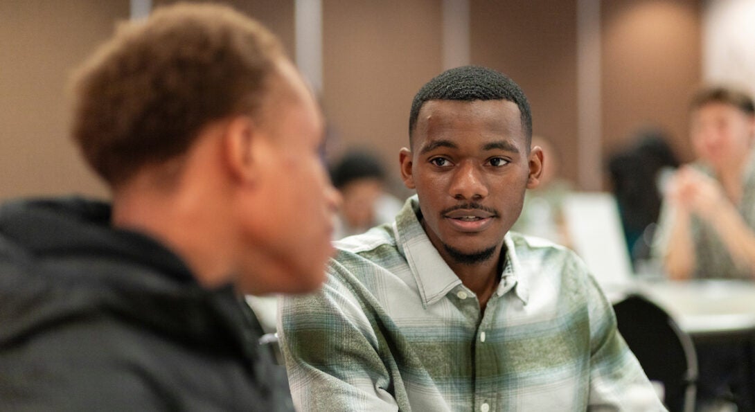 Integrated Health Studies student Emmanuel sits in a green and white button up shirt and talks to fellow student at event.