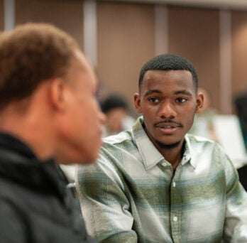 Integrated Health Studies student Emmanuel sits in a green and white button up shirt and talks to fellow student at event.
                  