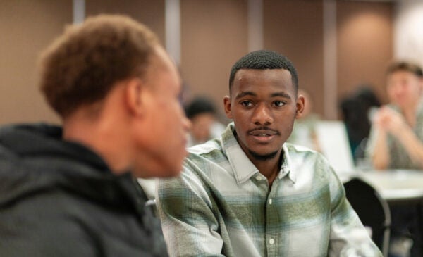 Integrated Health Studies student Emmanuel sits in a green and white button up shirt and talks to fellow student at event.