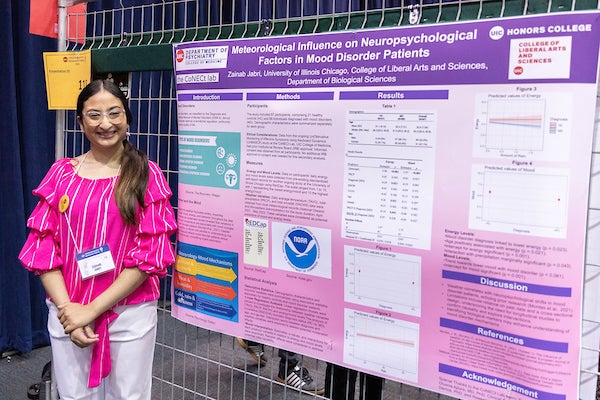 Young student wearing bright pink shirt stands next to her research poster at undergraduate research forum.