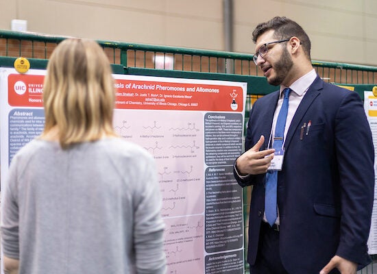 Young male student with short brown hair and brown beard with glasses and light skin describes his research project to event attendee.