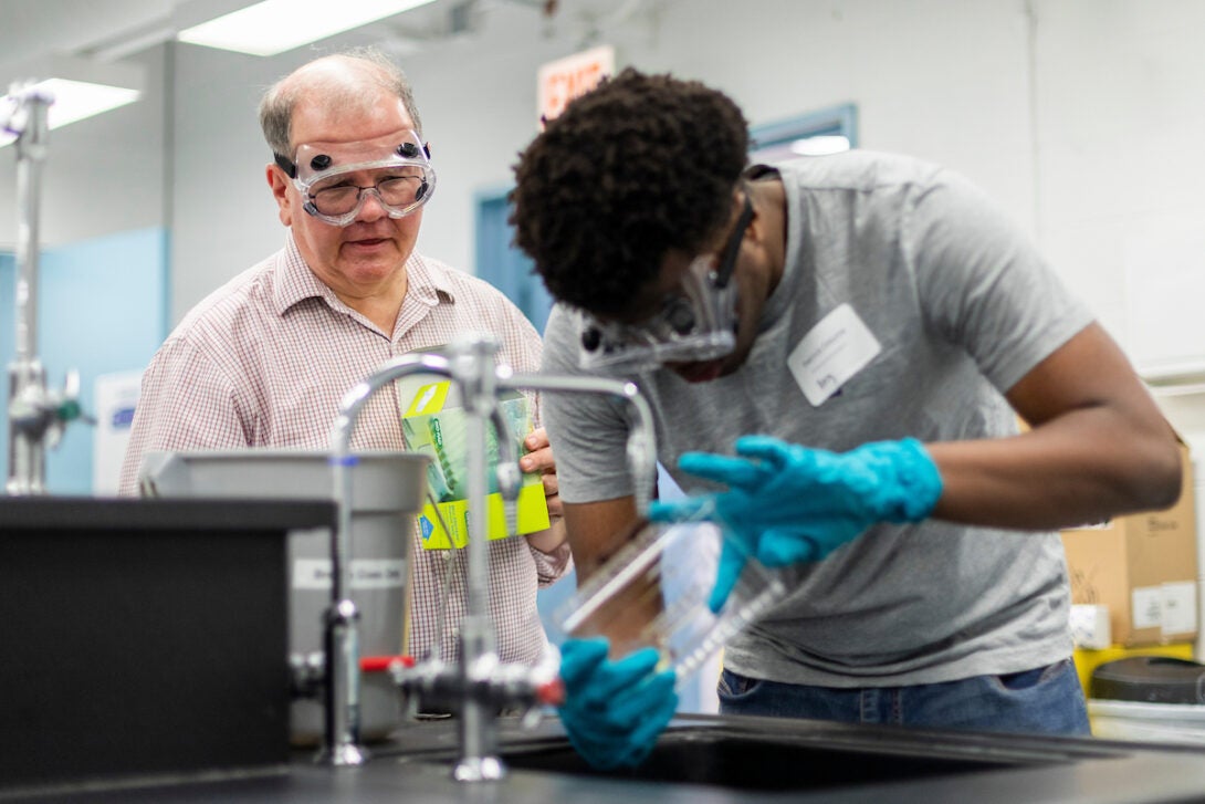 Young male black student pouring out science experiment into a sink while older white male professor overlooks and teaches.