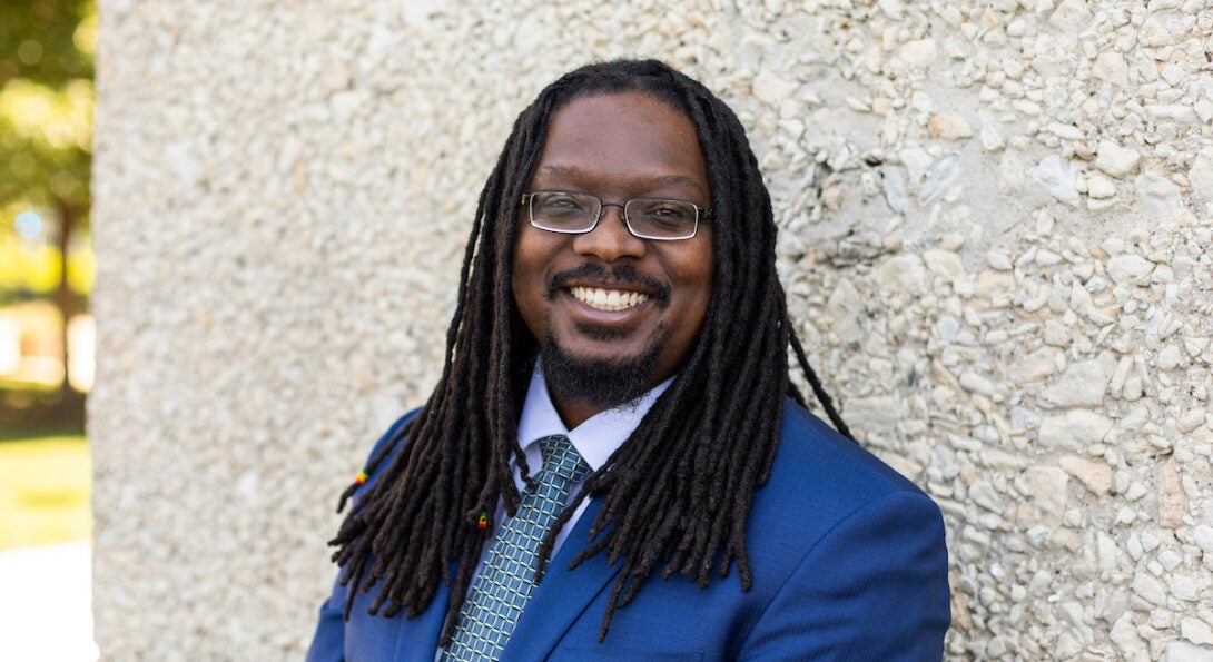 Director of IT Anthony Briscoe leans against a light pebble stone wall. He is a dark skinned black man with long dreadlocks and glasses and wears a light blue suit with a tie.