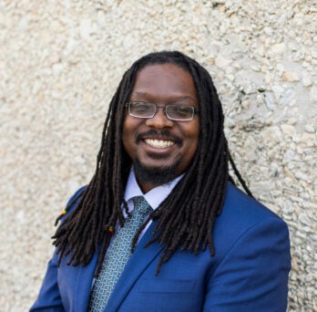 Director of IT Anthony Briscoe leans against a light pebble stone wall. He is a dark skinned black man with long dreadlocks and glasses and wears a light blue suit with a tie.
                  