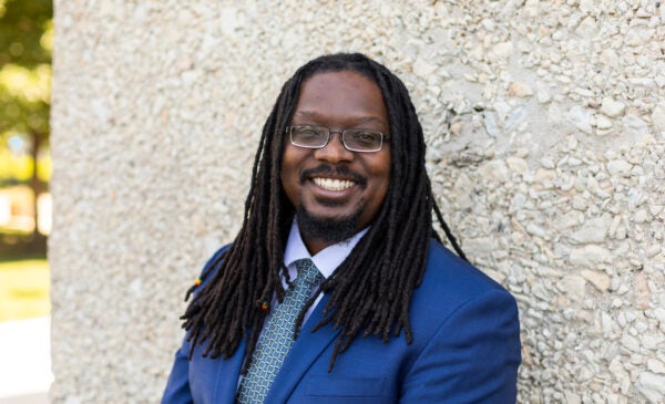 Director of IT Anthony Briscoe leans against a light pebble stone wall. He is a dark skinned black man with long dreadlocks and glasses and wears a light blue suit with a tie.