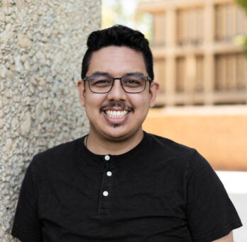 Alumni and current staff member Ivan Raya wears a black t-shirt, wireframe glasses, and has his dark hair cut short.
                  