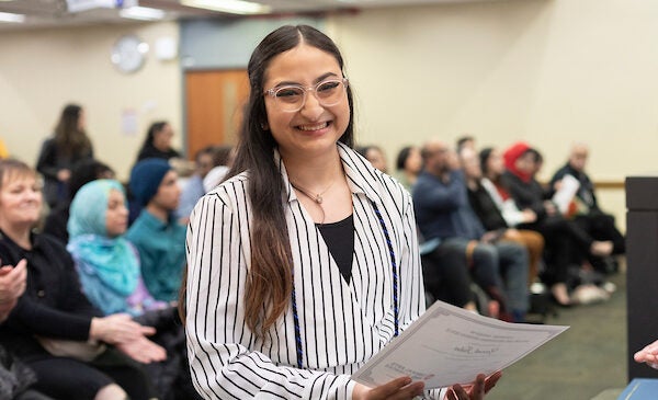 Female student with long brown hair and glasses holds her scholarship award and smiles.