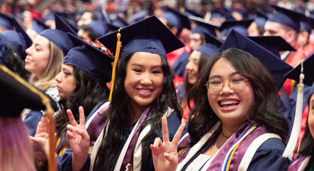 Two female students smile and show peace signs on their hands at commencement ceremony.