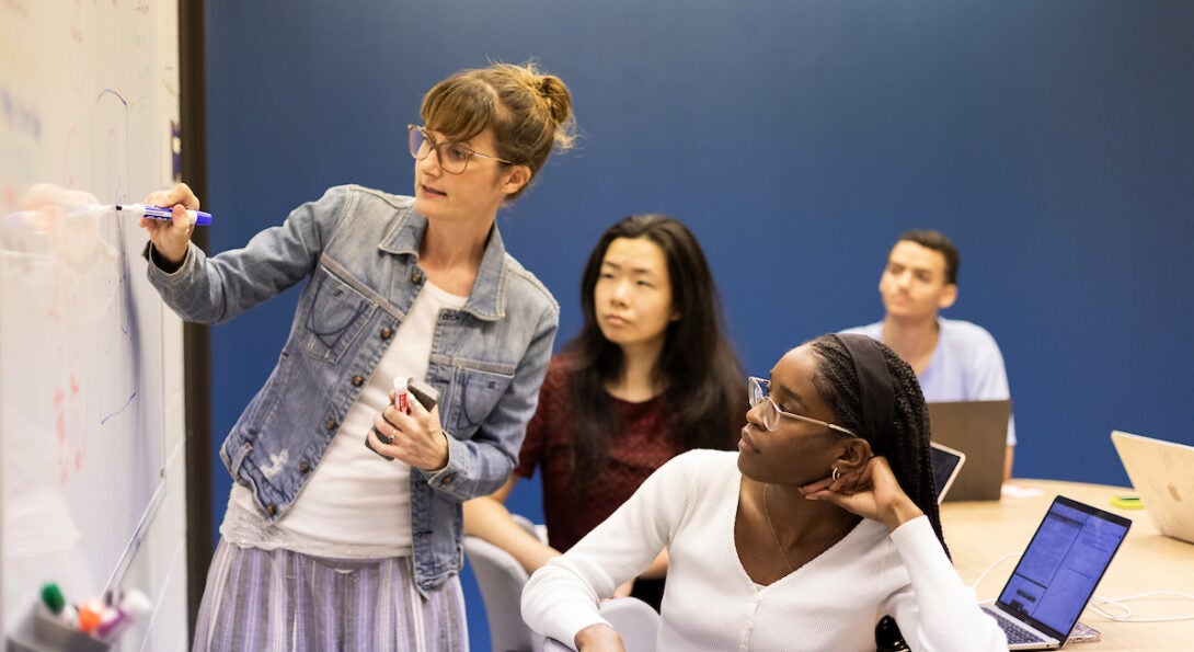 Professor writes on whiteboard in front of a classroom of diverse students.