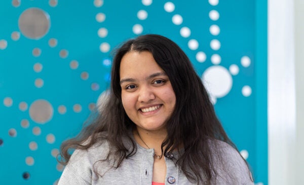 Young female student smiling in front of bright blue wall.