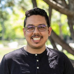 Man smiles and stands underneath tree on a sunny day. He is wearing glasses and a black tee shirt.