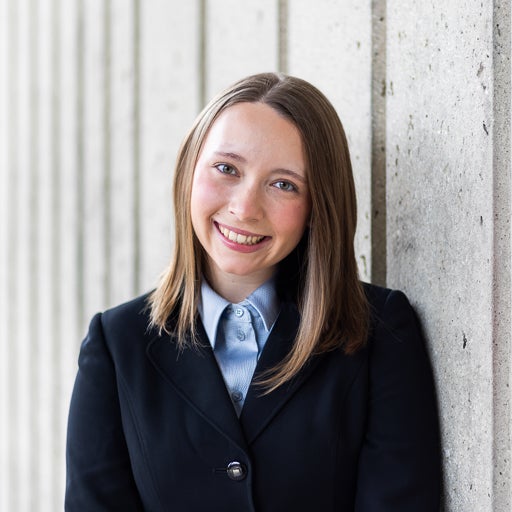 Student smiles against gray stone wall with black blazer and blue button up shirt