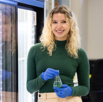 Biological Sciences student Iza Kopez in green sweater wearing blue rubber gloves in lab on UIC's campus
                  