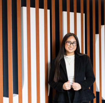 UIC Student Samantha Bravo standing in front of wooden logo sign wearing black blazer and white shirt
                  