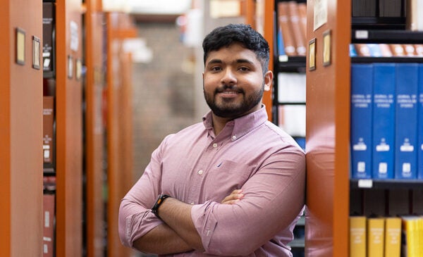 Student stands in library with arms crossed across chest. He is wearing a pink button up shirt, has short dark hair and a mustache and beard.